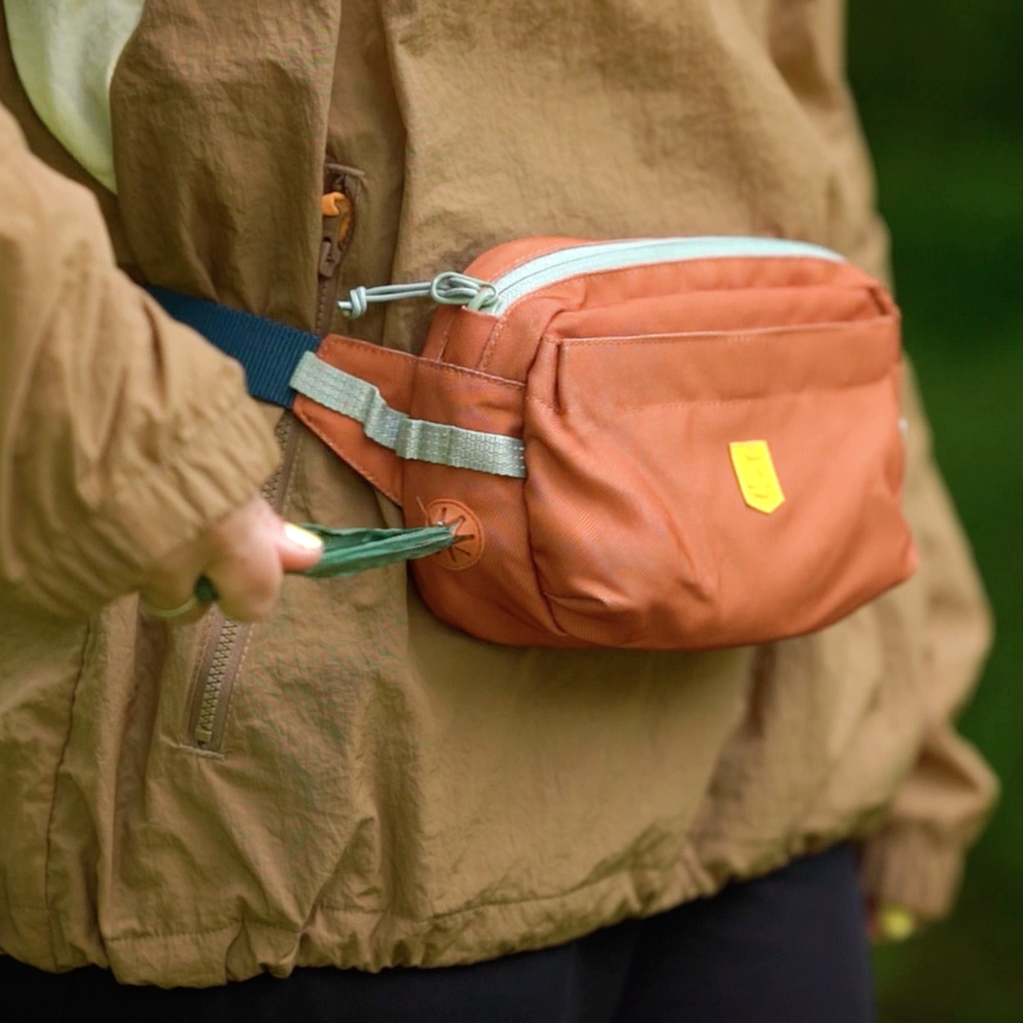 Person holding a phone and wearing an orange fanny pack over a brown jacket outdoors.