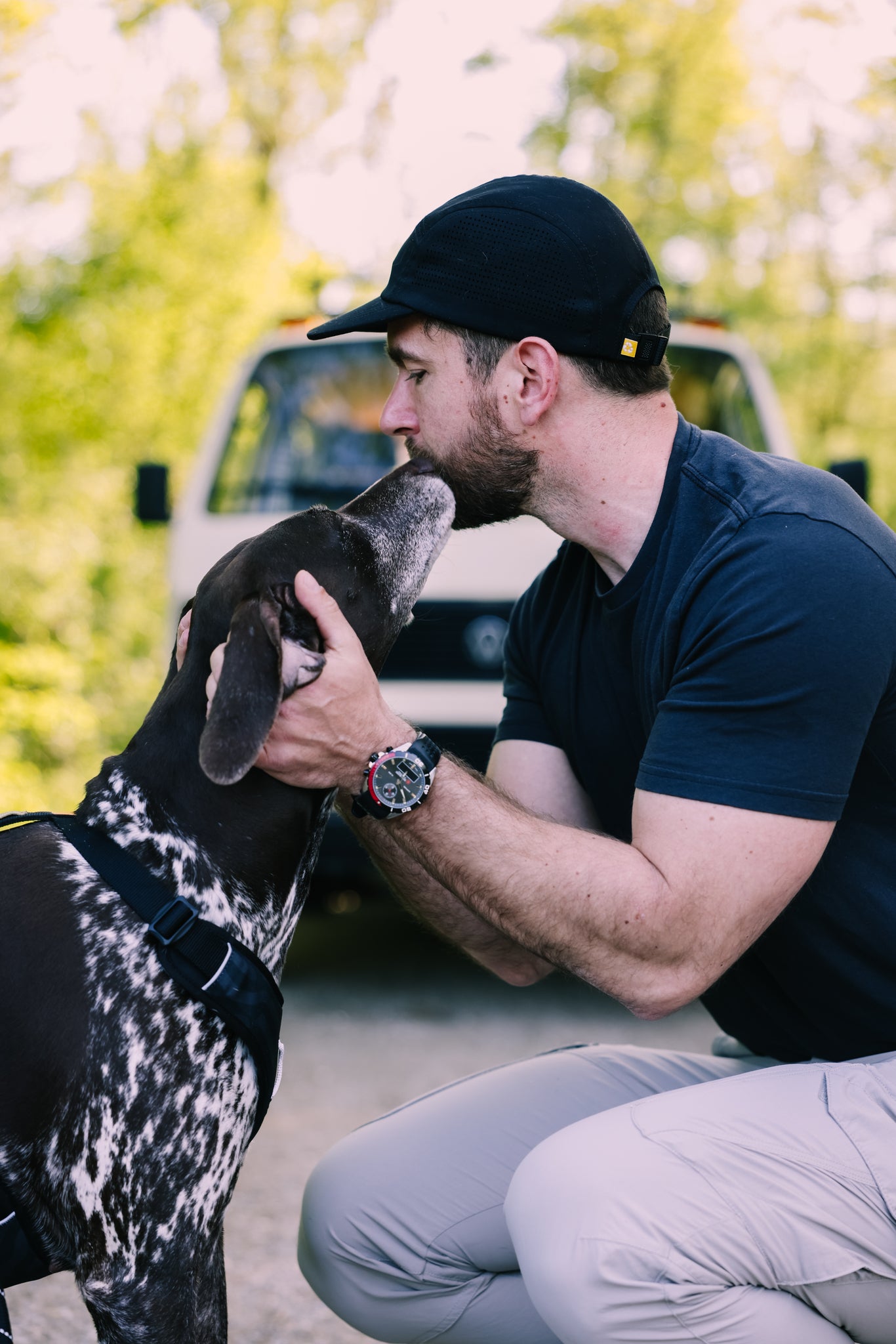A man wearing the Alpha 360 Tech Cap Black kneels outdoors, gently holding and kissing the head of a black and white dog, with a white vehicle and greenery blurred in the background.