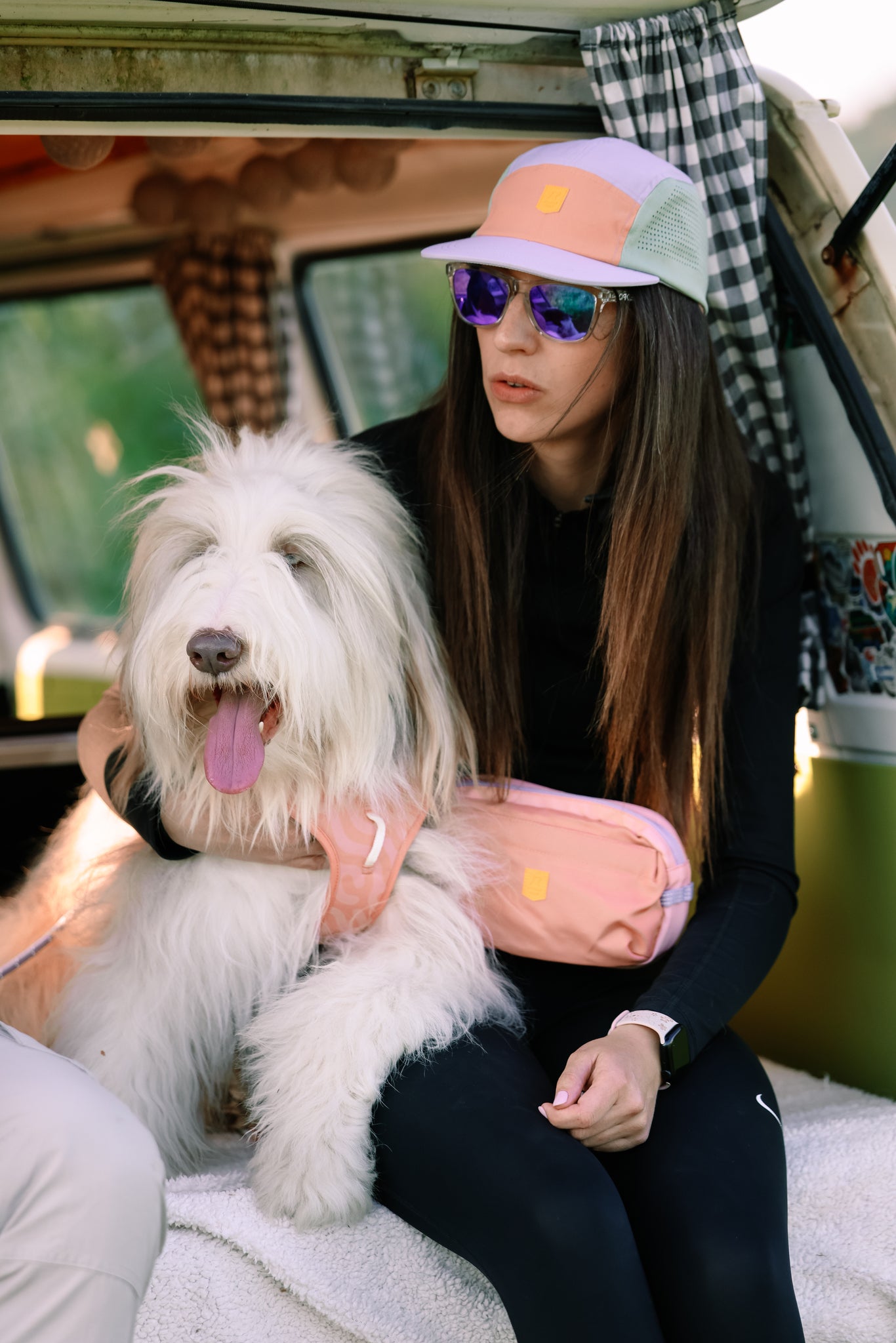 A woman in sunglasses wears the Alpha 360 Tech Cap Lavender and black clothing, sitting in a van with curtains and patterns. She holds a fluffy white dog with its tongue out and sports a pink fanny pack.
