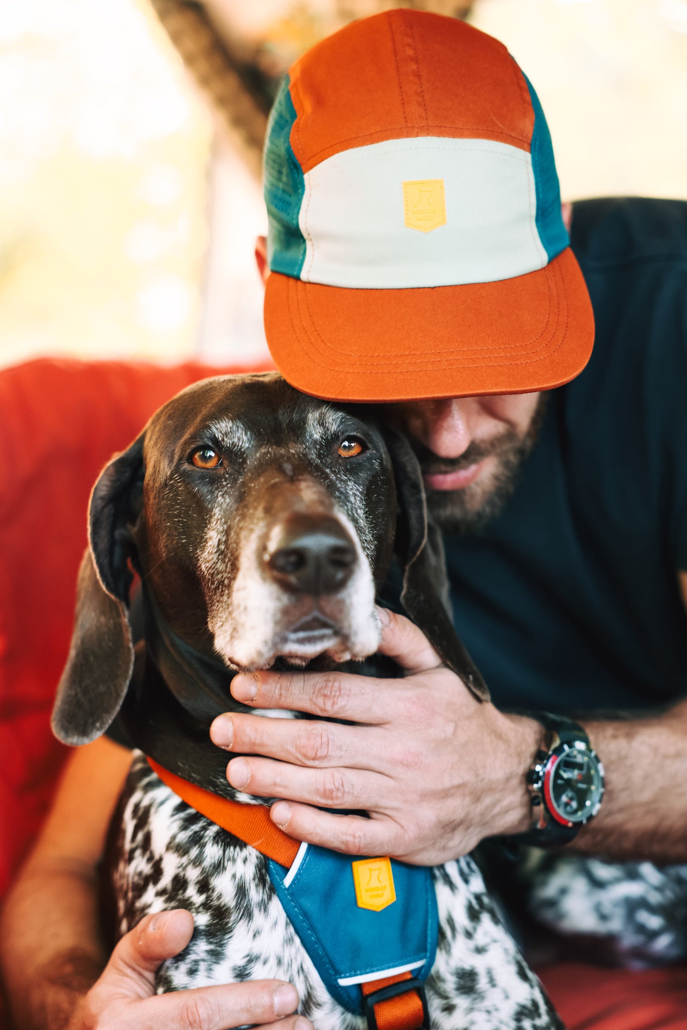 A man in an Alpha 360 Tech Cap Terracotta hugs a large black-and-white dog, gently holding its face. The cap partially hides his face, and both appear calm and close.