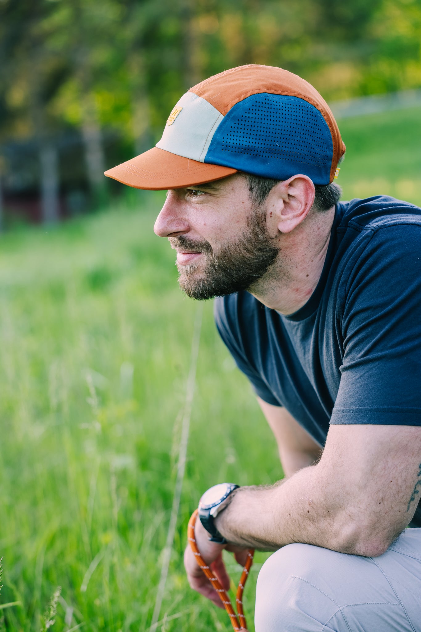 A bearded man wearing the Alpha 360 Tech Cap Terracotta and a dark t-shirt crouches in a grassy field, gazing thoughtfully to the left. Blurred trees and greenery fill the background.