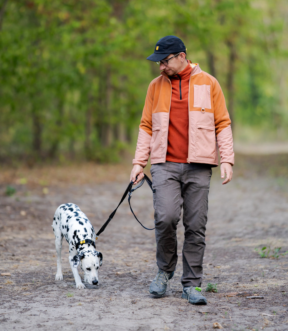 A man in a jacket and cap walks a Dalmatian on a leash through a wooded path.