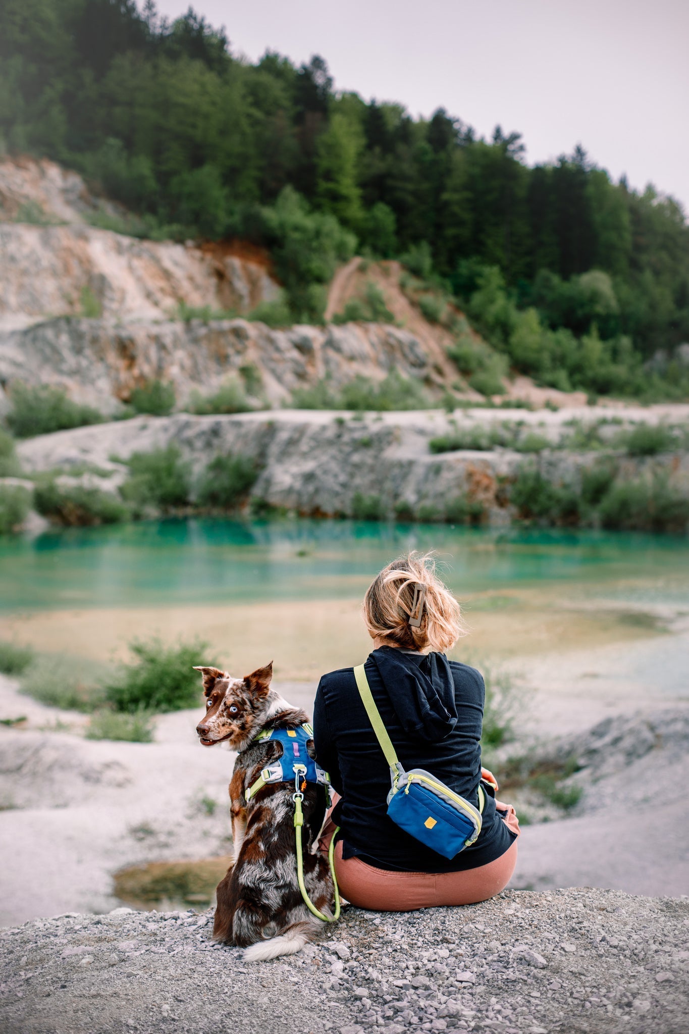 A woman with an Alpha 360 Hip Pack Deep Teal sits on a rock beside a dog, both facing a turquoise lake with rocky terrain and trees; the dog looks back at the camera.