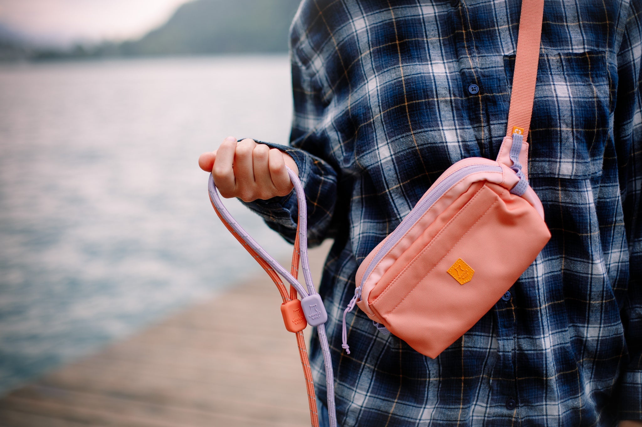 A person in a blue plaid shirt stands on a dock by the water, holding a pink leash and wearing the Alpha 360 Hip Pack in Salmon Pink. The background is slightly blurred, showing water and greenery in the distance.