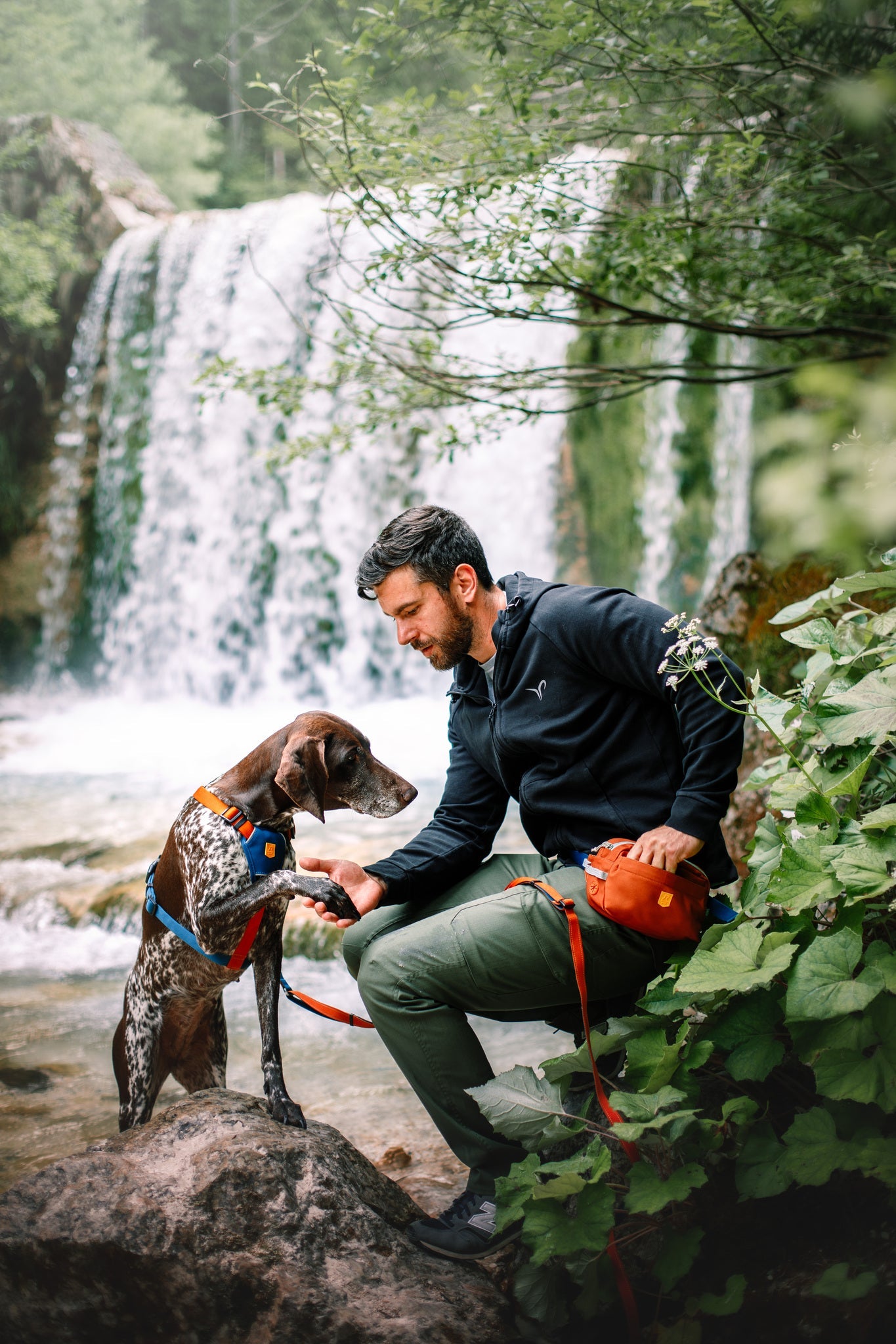 A man smiles and kneels next to his dog by a lush waterfall, wearing the Alpha 360 Hip Pack Terracotta Mix. Green foliage surrounds them as they relax outdoors with the hip pack visible around his waist.