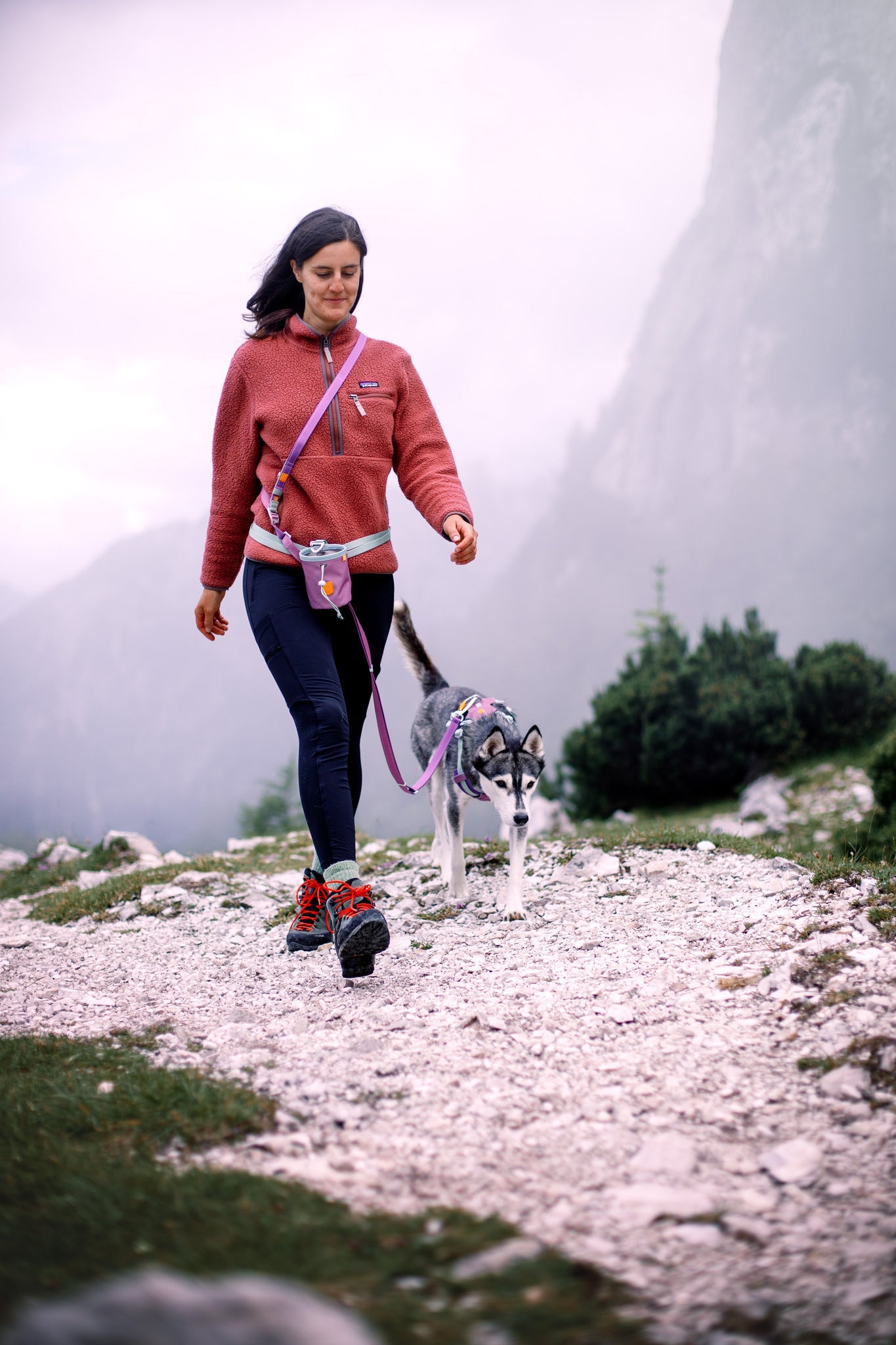 A woman in a red sweater walks a husky puppy on a rocky mountain trail, carrying the Alpha 360 Dog Snack Bag Mauve Mix, surrounded by misty mountains and lush greenery.
