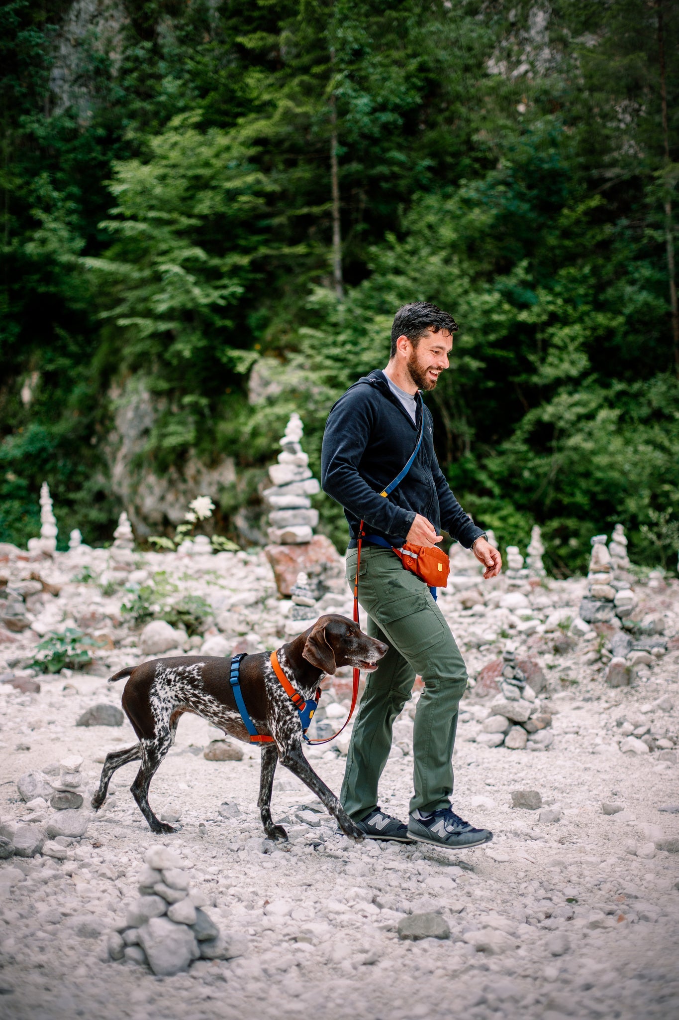 A man in a black sweater and green pants walks his brown and white dog, wearing a Color Block Dog Harness Lime Mix, on a leash along a rocky path with stacked stone piles and green trees in the background.