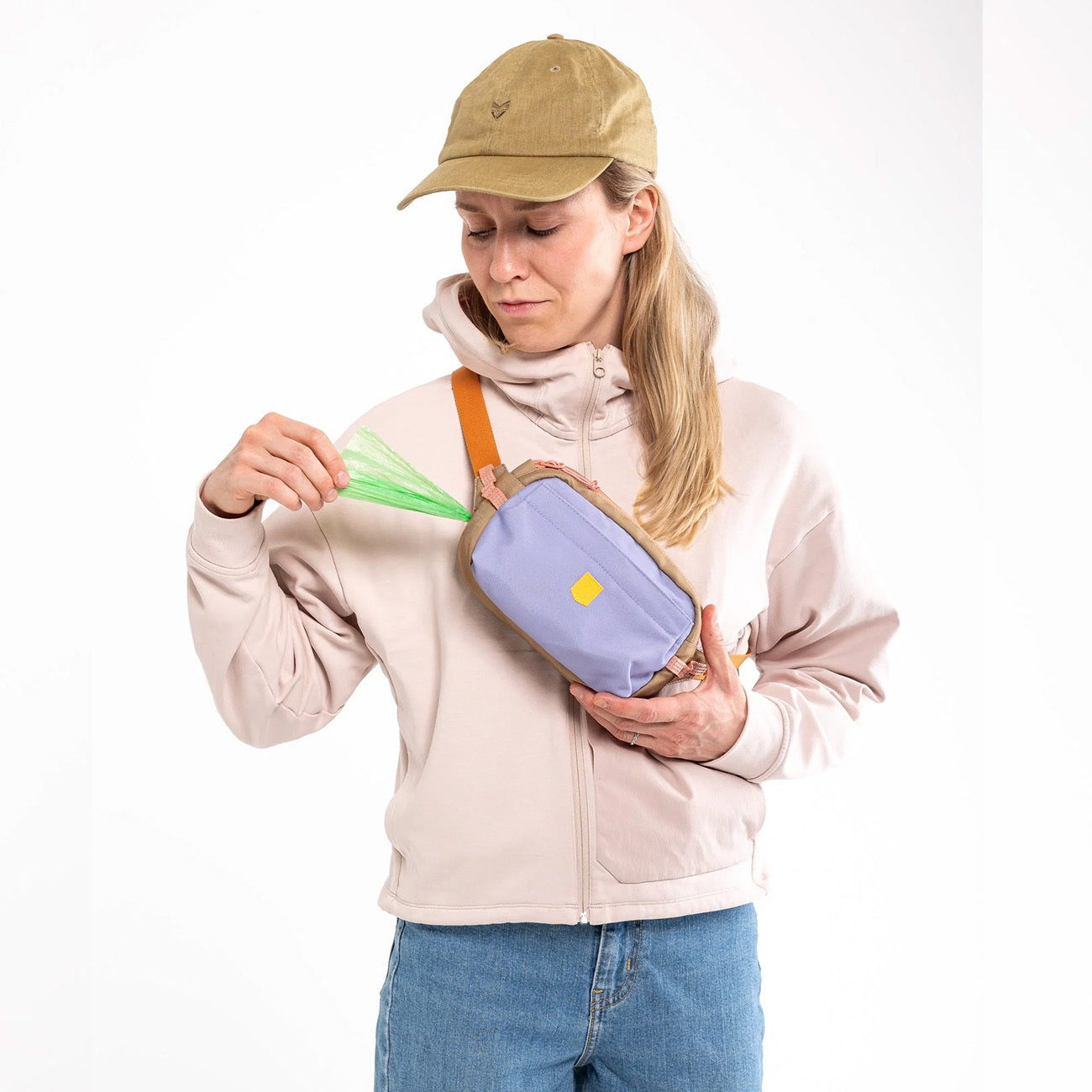 Woman in a beige hoodie and cap puts a green item into a small purple crossbody bag.