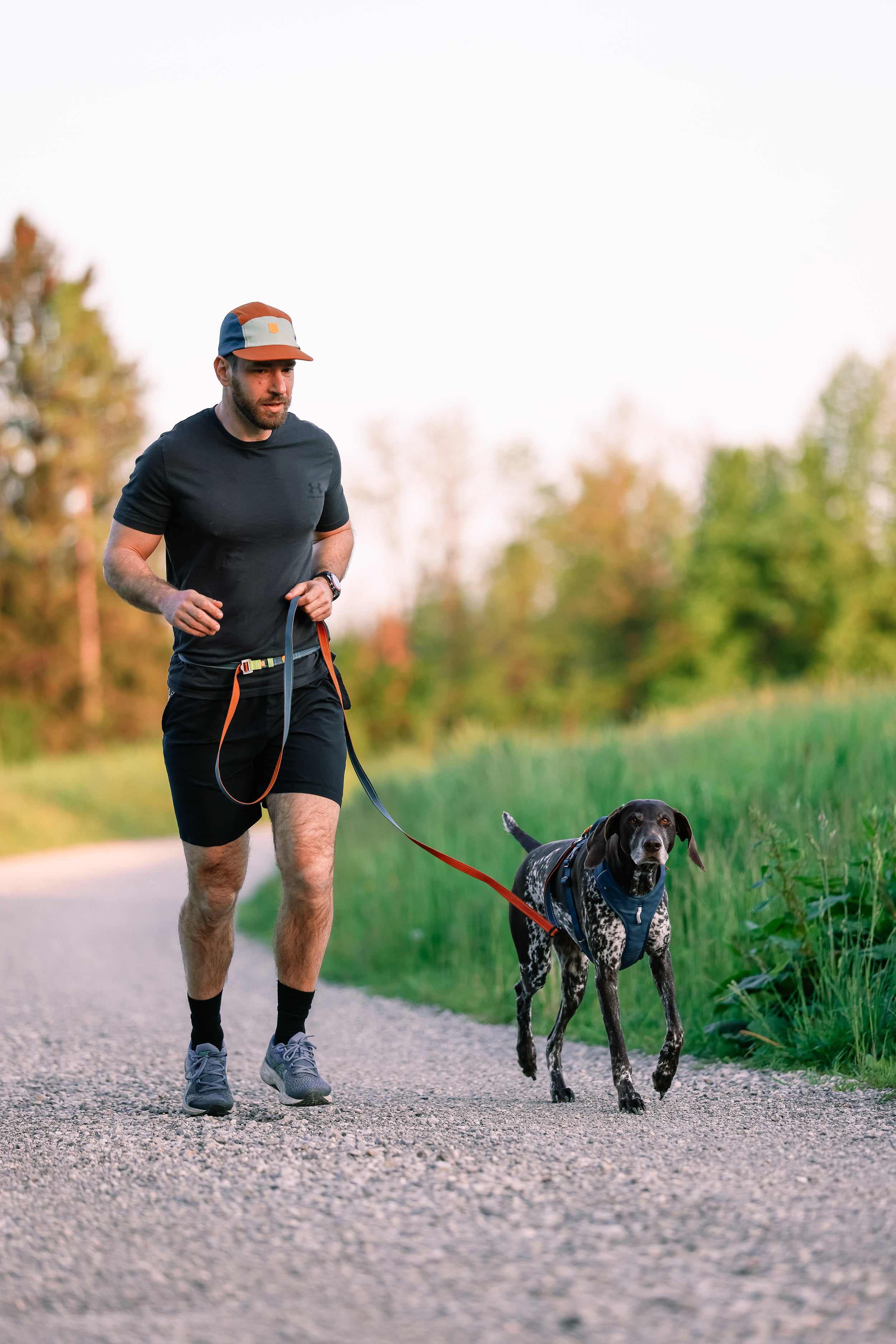 Man jogging on a gravel path with a black and white dog on a leash, surrounded by greenery.