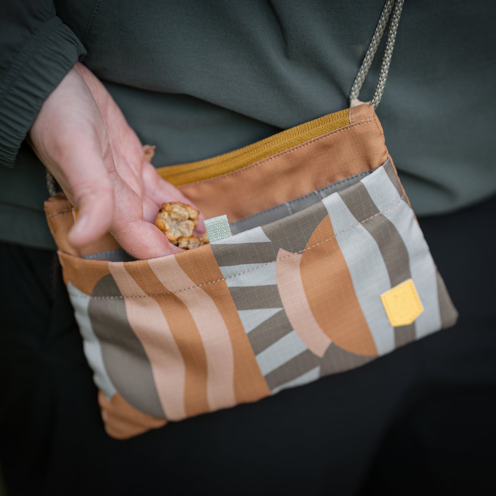 A hand takes a granola bar from a small, patterned fabric pouch with a zipper and shoulder strap.