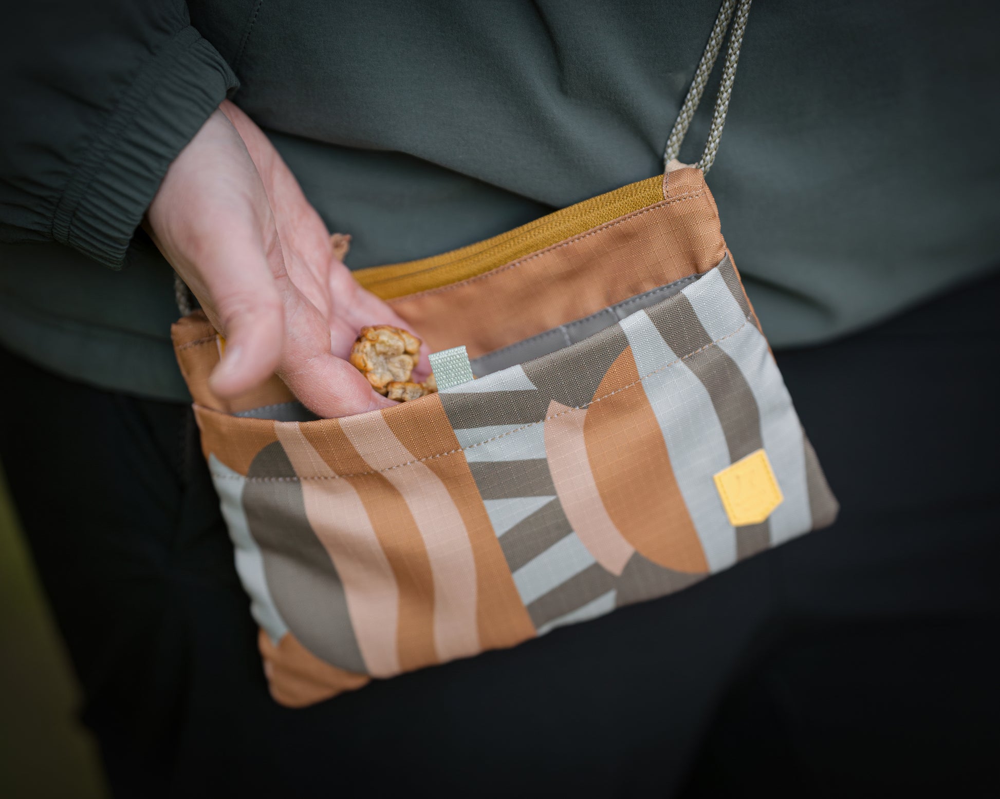 A hand takes a granola bar from a small, patterned fabric pouch with a zipper and shoulder strap.