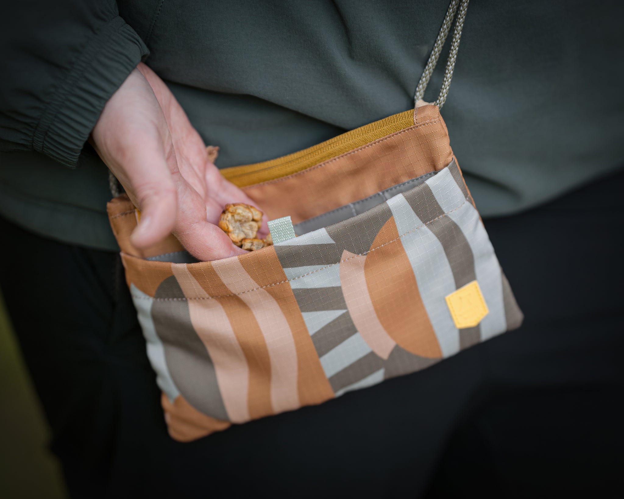 A hand takes a granola bar from a small, patterned fabric pouch with a zipper and shoulder strap.