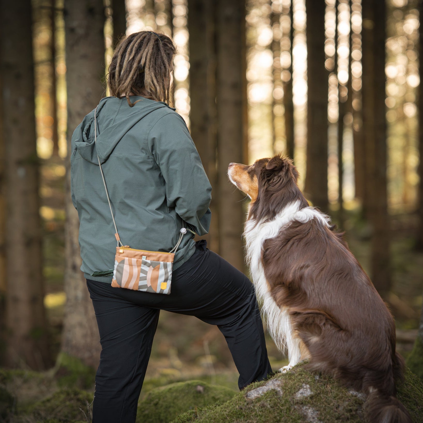 Person with dreadlocks and green jacket stands in forest with brown and white dog looking up at them.