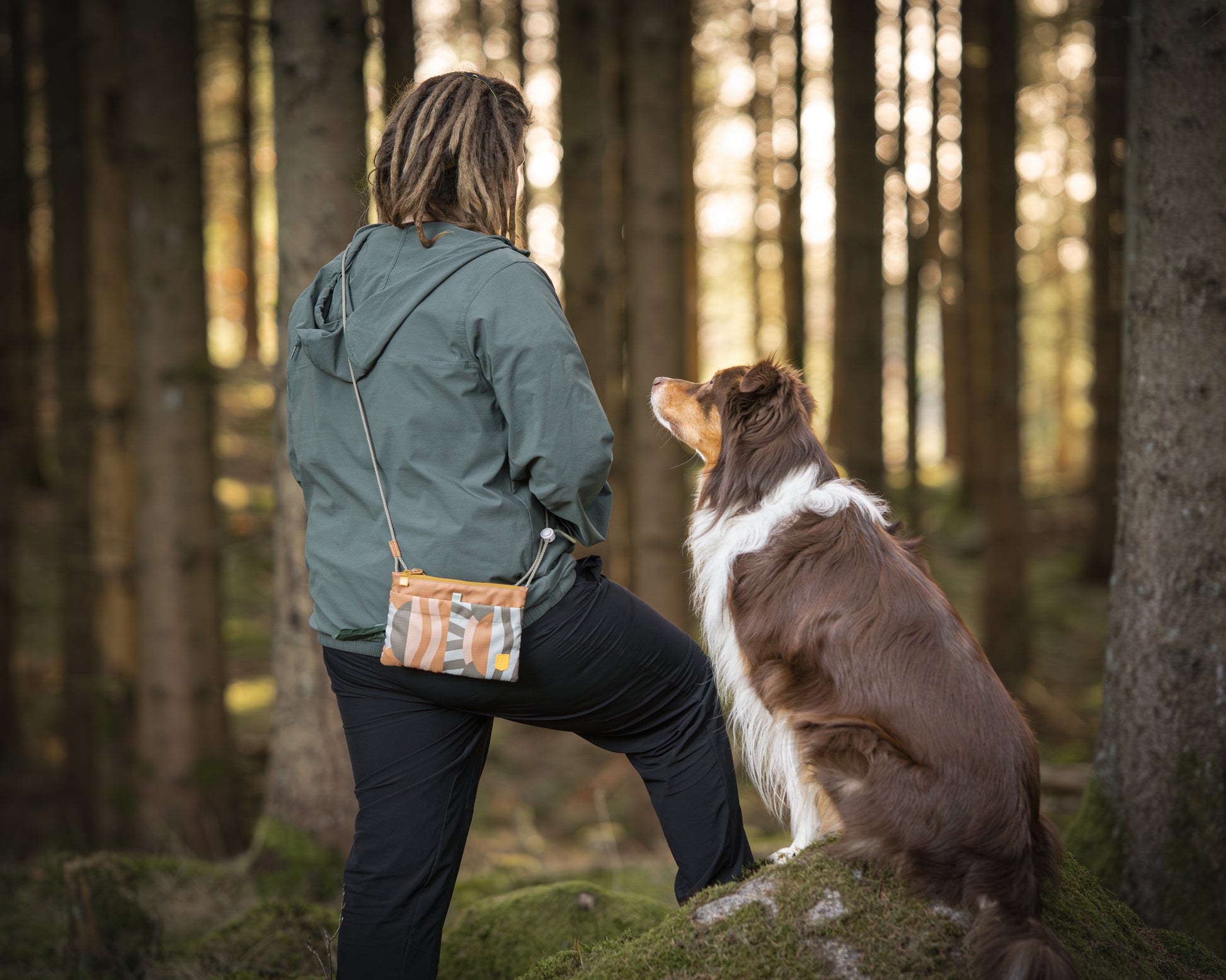 Person with dreadlocks and green jacket stands in forest with brown and white dog looking up at them.