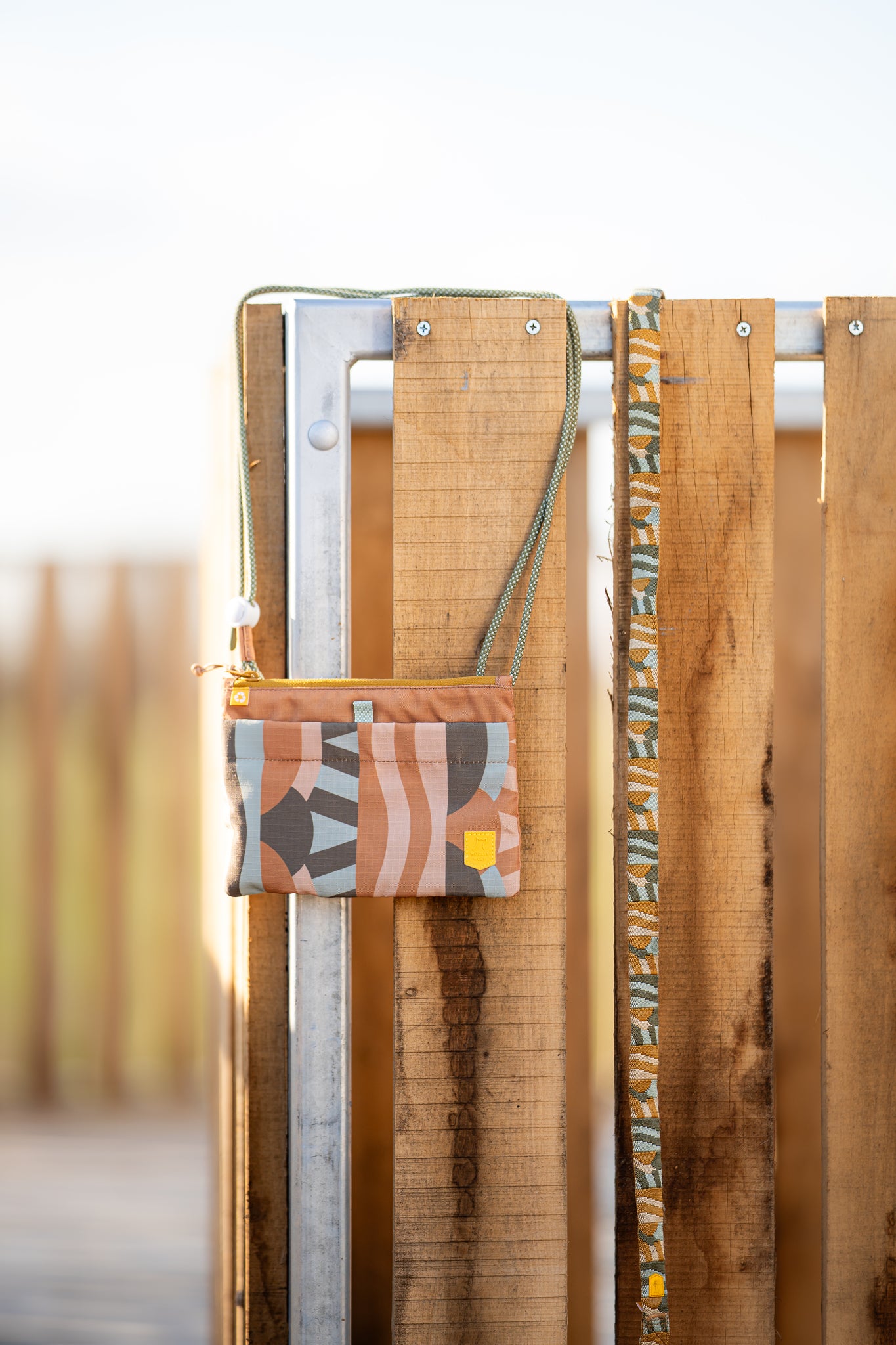 A small, patterned pouch with a strap hangs on a wooden and metal fence in bright sunlight.