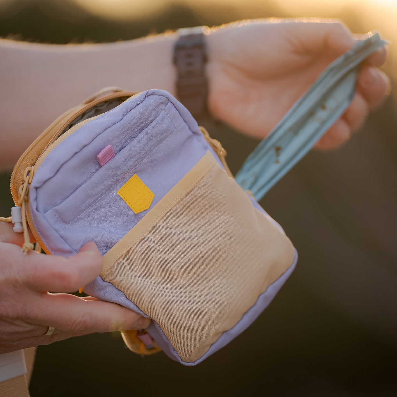 A person holds a small pastel crossbody bag and pulls out a teal face mask at sunset.