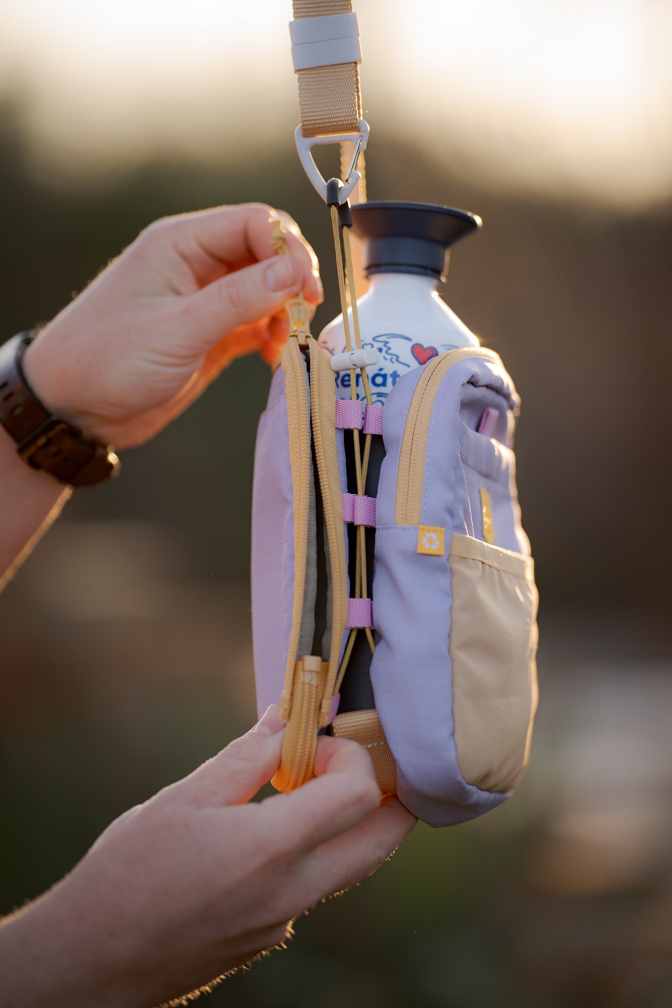 Person unzipping a small pastel bag with a water bottle inside, outdoors in soft sunlight.
