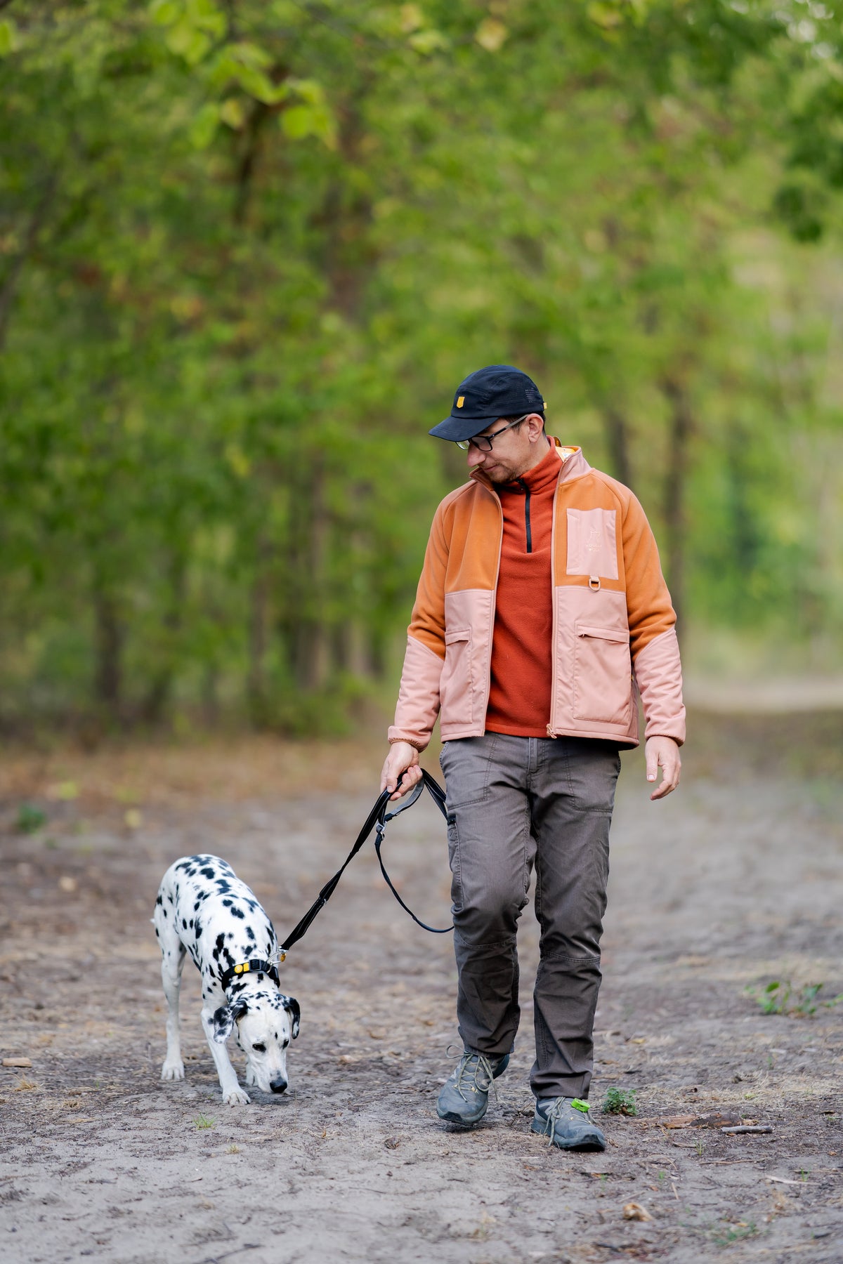 A man in a jacket and cap walks a Dalmatian on a leash through a wooded path.