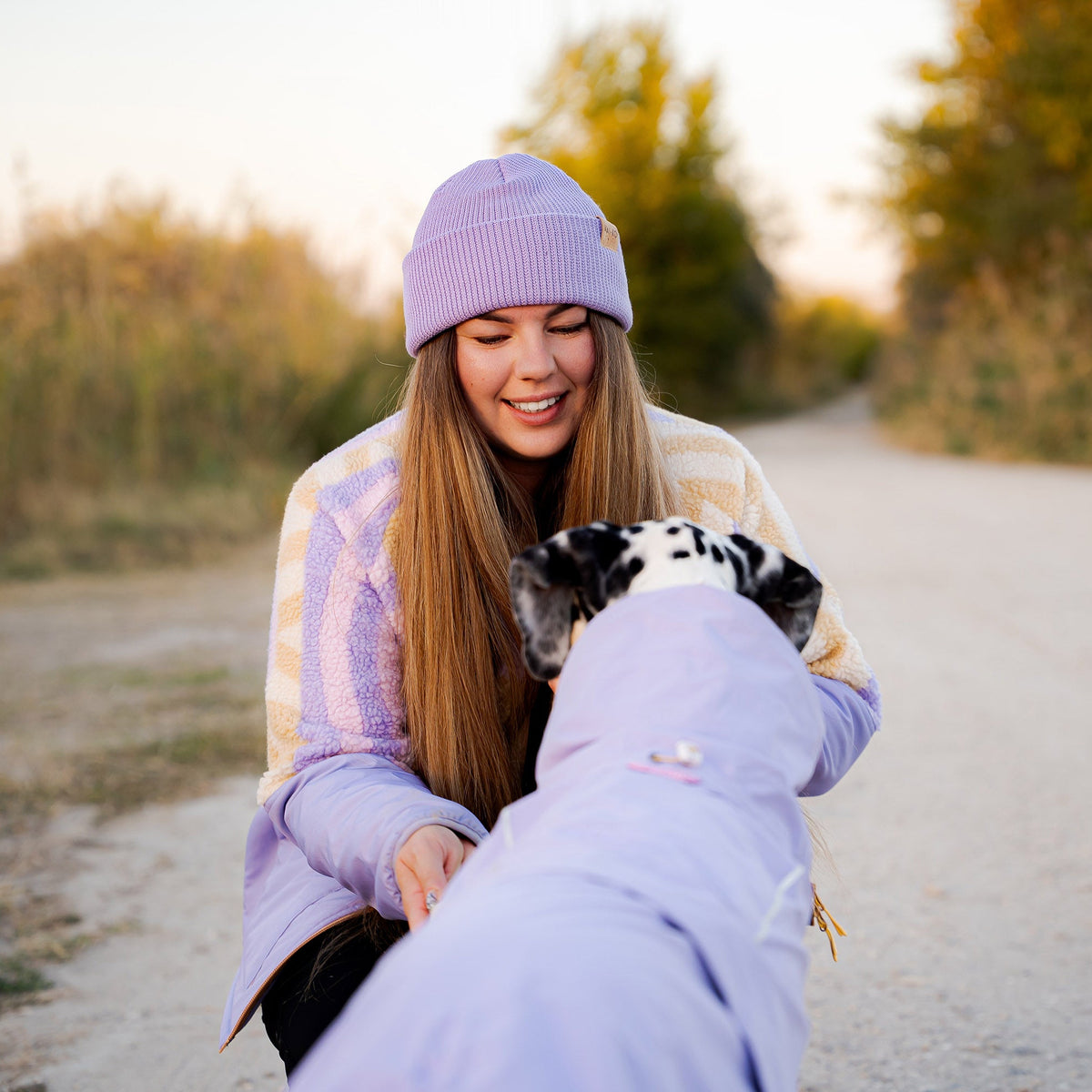 A young woman in a lavender beanie and jacket smiles at a dalmatian wearing a lavender coat.