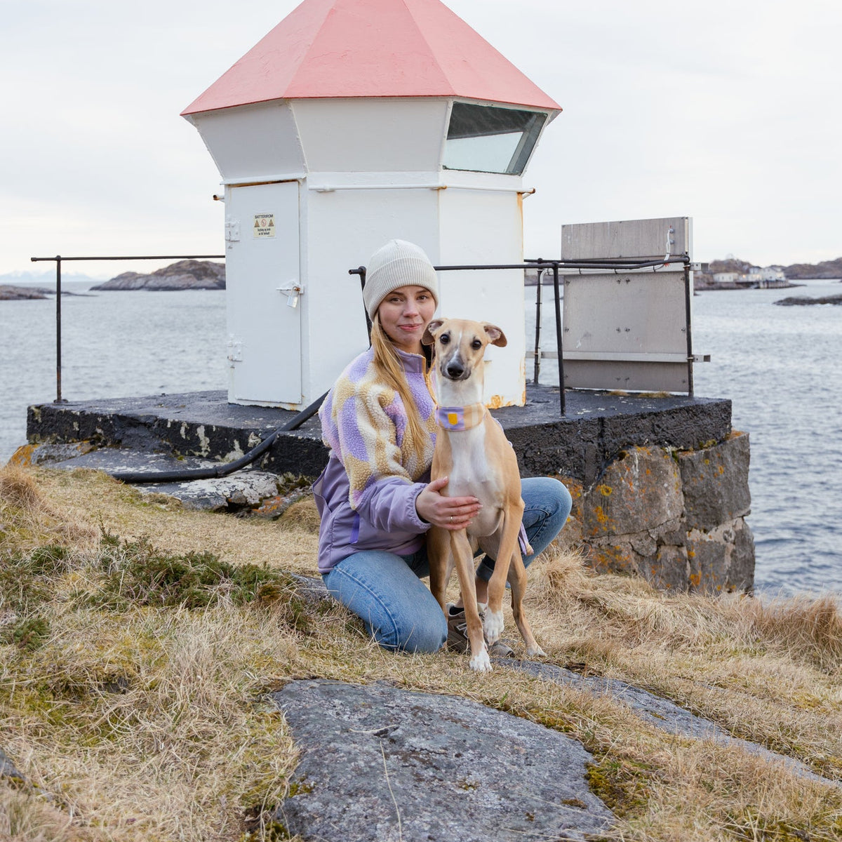Woman in a beanie kneels beside a dog on grass near a small lighthouse by the water.