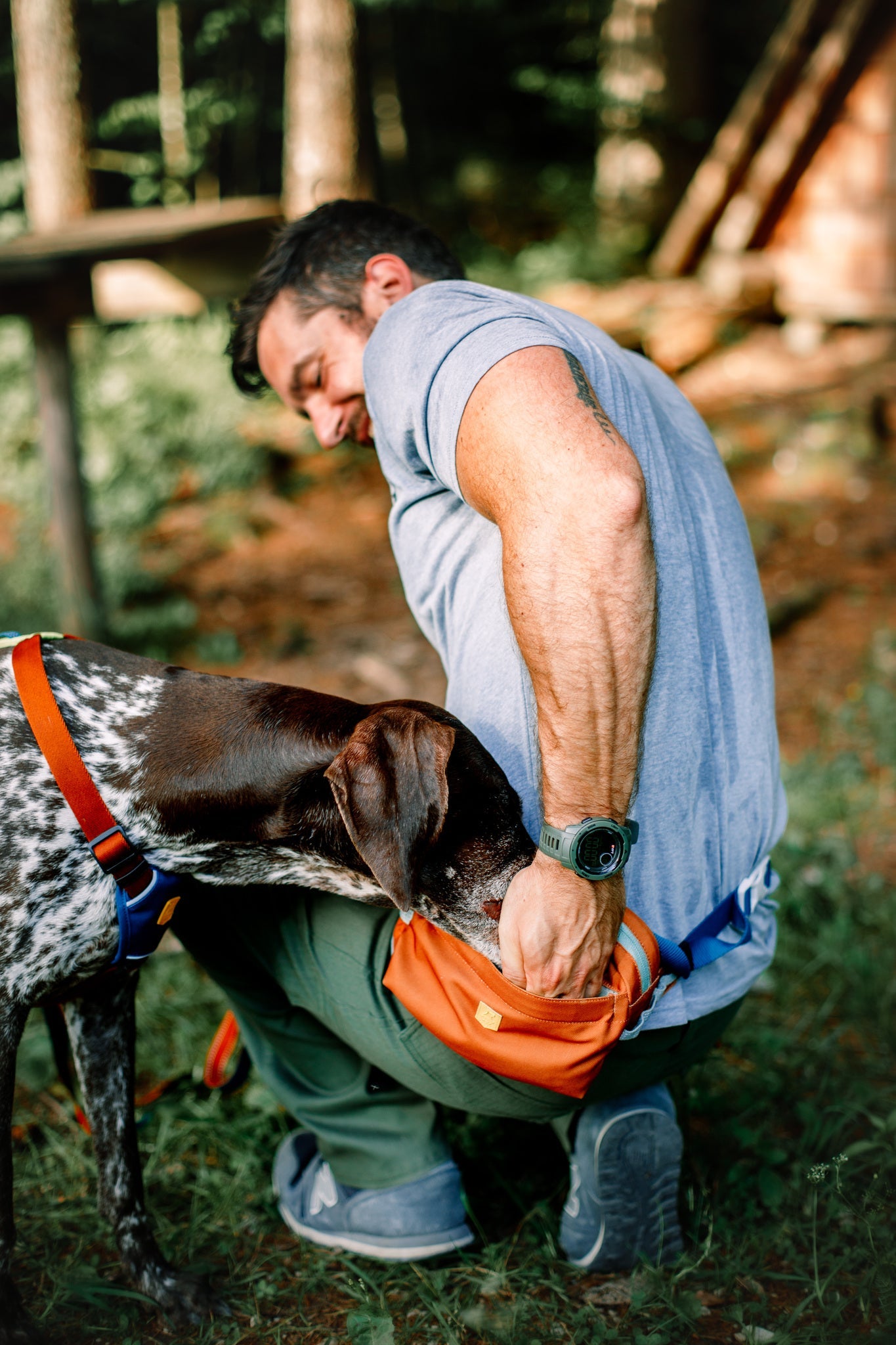 A man kneels outdoors, reaching into his Alpha 360 Hip Pack Terracotta Mix, while a brown and white dog with a red harness sniffs inside. Sunlight filters through the surrounding trees.
