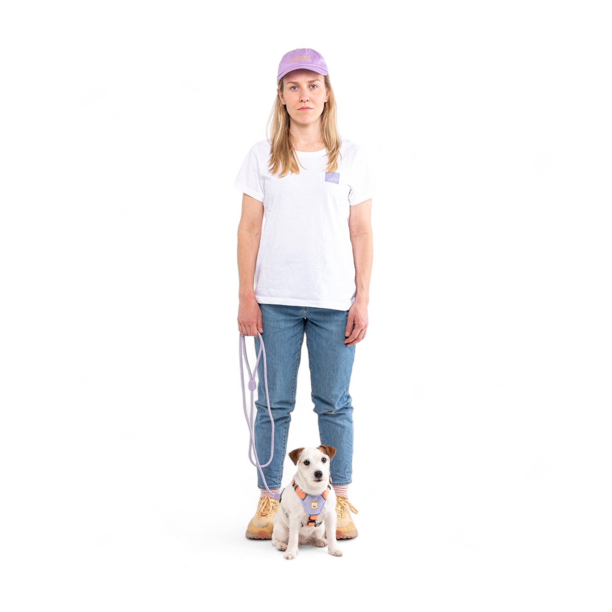 Woman in a lilac cap and jeans holds a leash beside a Jack Russell terrier wearing a harness.